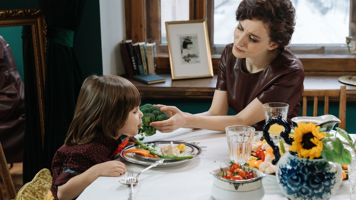 A caring mother helps her child eat healthy vegetables at a dining table, promoting family bonding.